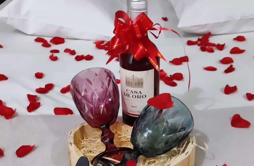 Cama de casal com vinho, taças e pétalas de rosas