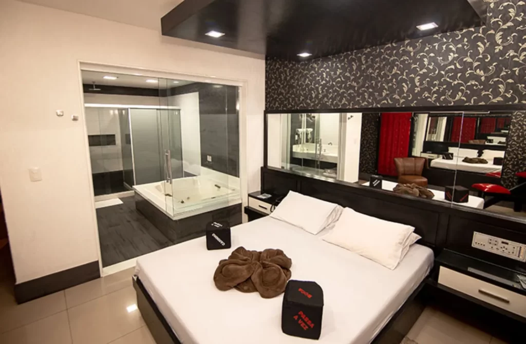 Quarto de motel com cama de casal, espelhos ao redor e banheiro ao lado com banheira