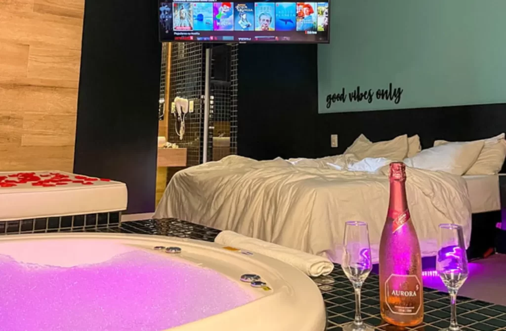 Quarto de motel com cama de casal, champagne, banheira, TV e rosas ao redor