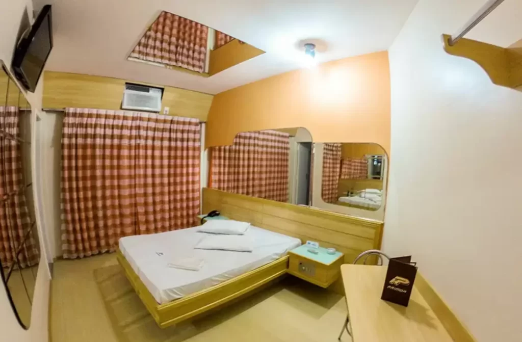 Quarto do hotel com cama de casal, TV, cortina, mesa com cadeiras e espelhos ao redor