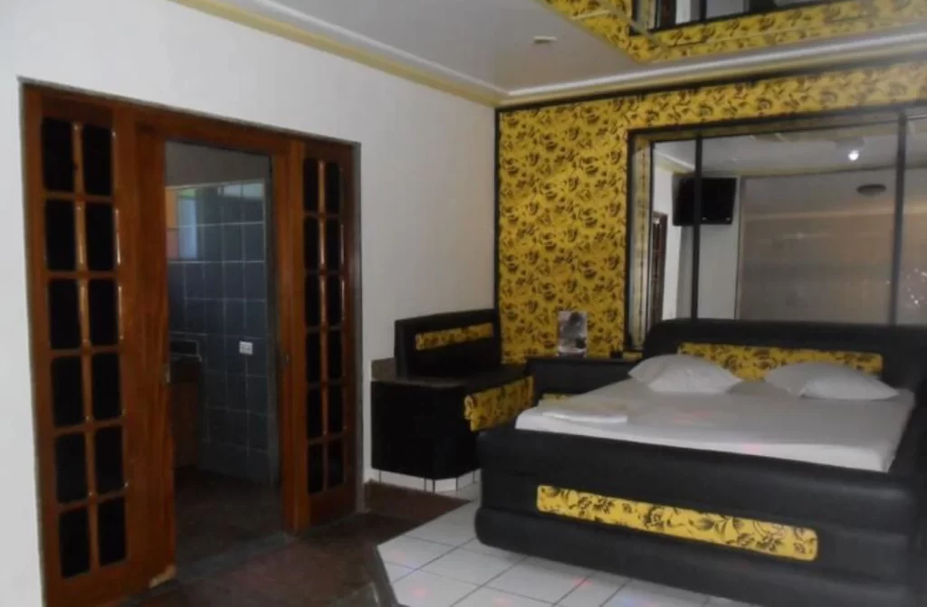 Quarto de hotel com paredes amarelo e preto, cama de casal, espelhos e banheiro do lado