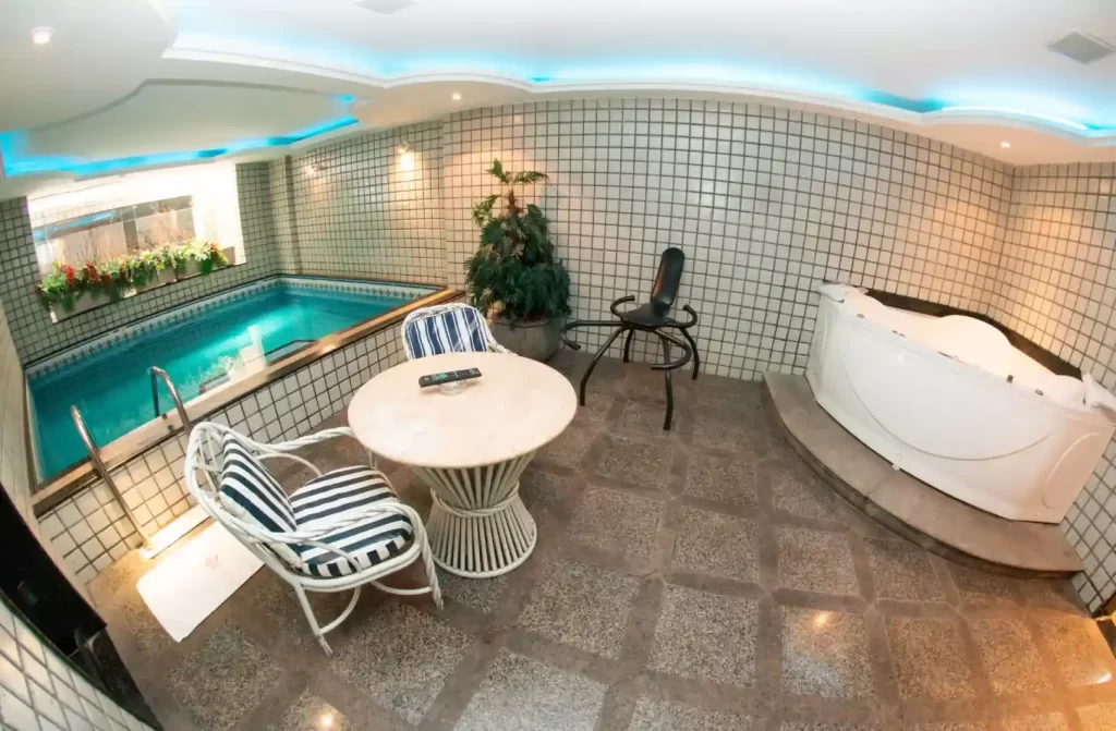 Área de piscina de um motel com mesa, cadeira, banheira, piscina e plantas decorativas